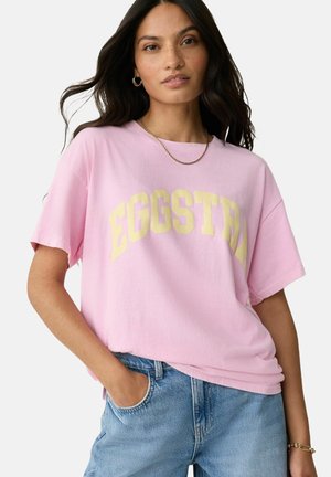 Pink cotton t-shirt featuring the printed word "EGGSTRA" in yellow. Relaxed fit with short sleeves and a round neckline. Minimalist design.