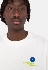 White sweatshirt with a round blue patch and vibrant green text "GRIP ROOTS ROCKS." Features a classic crew neckline and soft texture.