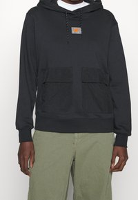 Svart huvtröja med stora framfickor, grå logotyp-patch, dragsko på huvan och ribbade muddarna. Har en mjuk tygkänsla.