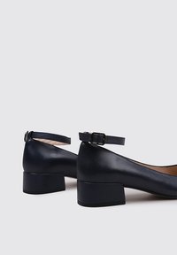 Zapatos de tacón bloque de cuero azul marino con correa en el tobillo, puntera redondeada, textura suave y diseño minimalista. Acabados con una pequeña hebilla metálica.
