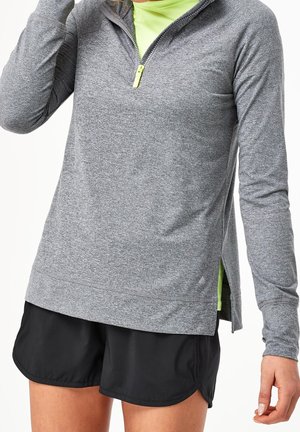 Femme portant un haut gris à manches longues zippé sur un t-shirt vert fluo et un short de sport noir.