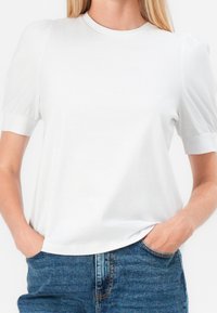 Top blanco de manga corta con mangas abullonadas, escote redondo y textura suave. Combinado con jeans de mezclilla azules, exhibiendo una apariencia casual.