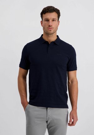 Mann steht mit den Händen in den Taschen, trägt ein marineblaues Polohemd mit kurzen Ärmeln und hellgraue Hosen vor einem schlichten Hintergrund.