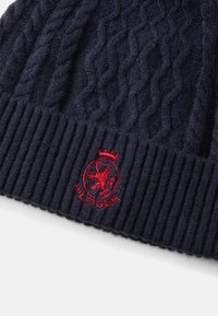 Navy gebreide beanie met een geribde manchet, voorzien van een verheven rode leeuw-embleem en een ingewikkeld kabelbreipatroon op het voornaamste deel.