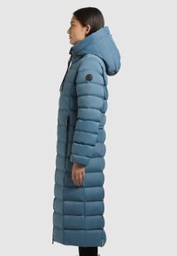 Kvinde i en teal lang pufferjakke med hætte, stående i sideprofil foran en ensartet lys baggrund.