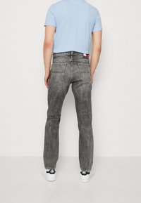 Vaqueros de mezclilla gris con acabado desgastado, cinco bolsillos y un pequeño parche con logo en la cintura. Combinados con una camiseta azul claro y zapatillas blancas.