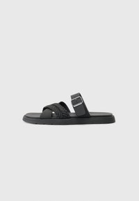Black sandal featuring braided straps, a flat sole, and a buckled ankle strap. Made from synthetic material with a smooth finish.