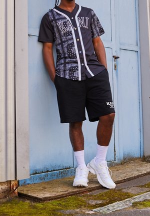 Man leunend tegen een blauwe deur, gekleed in een zwart patroonhemd, zwarte korte broek, witte sokken en witte sneakers buiten.