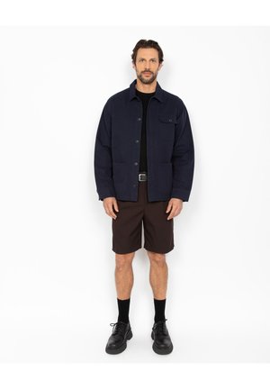 Homme portant une veste marine à boutons, une chemise noire, un short marron foncé, des chaussettes noires et des chaussures en cuir noir, debout contre un fond blanc.