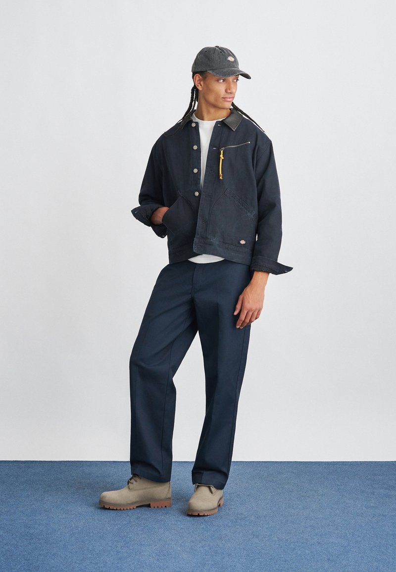 Young person with braided hair wearing a black cap, navy jacket with yellow zipper pull, white shirt, navy pants, and beige boots on blue floor.