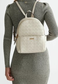 Zaino Liu Jo in pelle beige goffrata con logo dorato indossato sopra un vestito grigio a coste sulla schiena di una persona.