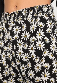 Tissu noir avec un imprimé de marguerites blanches et jaunes, présentant un détail de taille froncée. La texture générale semble douce et légère.