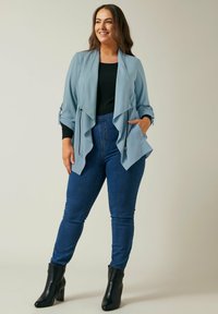 Vaaleansininen, virtaava takki, jossa on laskostettu etuosa ja käännetyt hihat, yhdistettynä tummansinisiin slim fit -farkkuihin ja mustiin nilkkureihin.