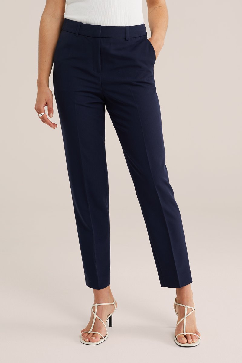Vrouw draagt marineblauwe, op maat gemaakte enkellange broek, witte sandalen met hak en bandjes, en een wit mouwloos topje, staand met één hand in de zak.