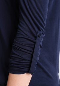 Top de manga larga de color azul marino con textura fruncida en la manga. Presenta un detalle de botón cerca del puño para mayor interés. Tela suave y elástica.