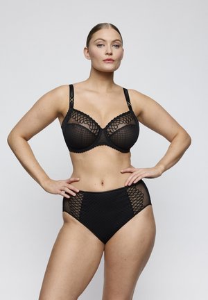 Woman with hair tied back wearing a matching black lace bra and high-waisted panties, standing with hands on hips against a plain background.