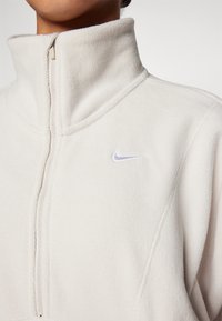 White fleece pullover with a high collar, half zip, and a small purple Nike logo embroidered on the chest. Soft texture and seamless design.