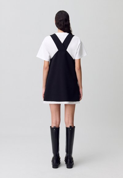 Vestito nero con una camicia bianca sotto, caratterizzato da un design senza maniche e spalline regolabili, abbinato a stivali neri al ginocchio.