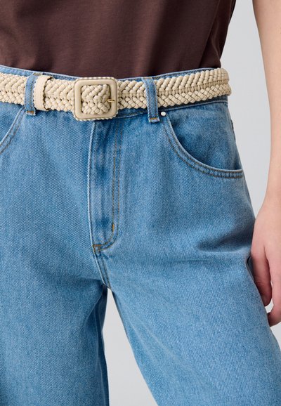 Cinturón trenzado beige con hebilla cuadrada, usado sobre unos jeans de mezclilla azul claro con diseño de cintura alta y un sutil patrón texturizado.