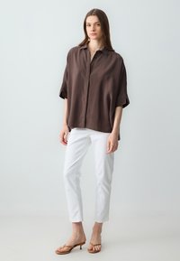 Braunes kurzärmliges Hemd mit Knöpfen, entspannte Passform, mit glatter Textur, kombiniert mit weißen Slim-Fit-Hosen und beigen Sandalen.