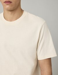 T-shirt beige à manches courtes en coton doux, avec un col rond et une texture lisse. Aucun motif ni accent visible.