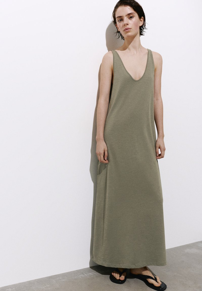Jeune femme aux cheveux courts et foncés, vêtue d'une robe maxi olive sans manches et de tongs noires, debout contre un mur blanc.