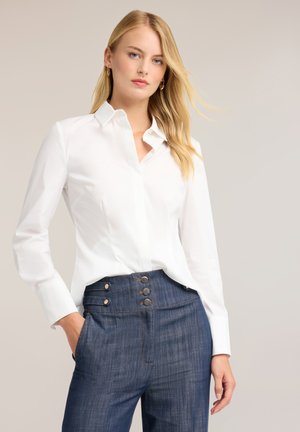 Junge Frau mit langen blonden Haaren, die ein tailliertes weißes Hemd und hochgeschnittene blaue Jeans mit Knopfdetails trägt.