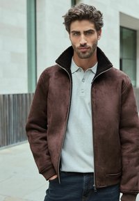 Jachetă din suede fals maro, cu guler căptușit cu sherpa, închidere cu fermoar și manșete cu dungi, purtată peste un polo texturat gri deschis și blugi negri.
