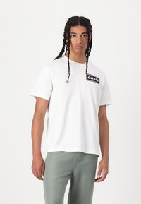 White cotton t-shirt with short sleeves and a round neckline, featuring a black rectangular "oasis" logo on the chest. Model wears light green pants.