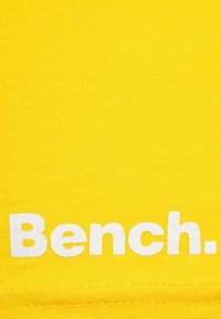Tela amarilla con una textura suave y el texto impreso en blanco "Bench." en la parte inferior, enfatizando el diseño de la marca.