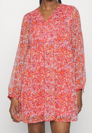 Kobieta w mini sukience z długim rękawem, w kwiatowy wzór w kolorach czerwonym, pomarańczowym, różowym i lawendowym, z dekoltem w serek.