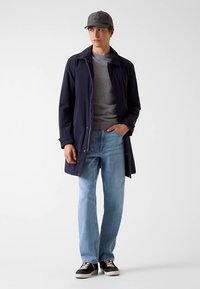 Veste imperméable bleu marine portée sur une chemise grise, associée à un jean bleu clair et des baskets noires. Le modèle porte une casquette grise.