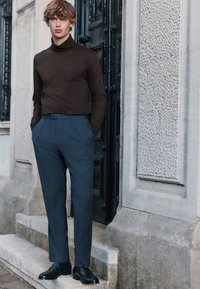 Brun polotröja, blå klänningsbyxor och svarta skor. Mjukt tyg med en figursydd design. Står mot en strukturerad vägg.