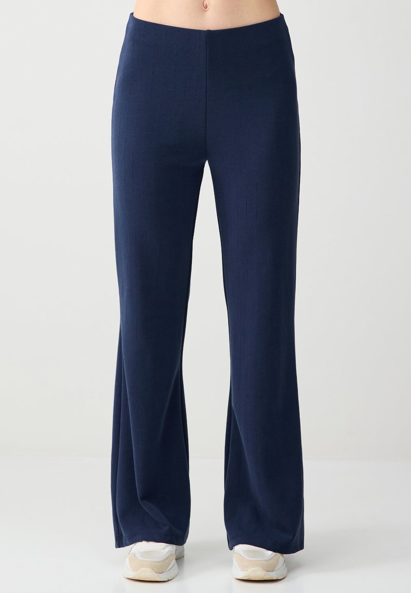 Marineblauwe geribbelde broek met brede pijpen, gedragen met witte sneakers, waarbij de buik en onderbenen zichtbaar zijn tegen een effen lichte achtergrond.