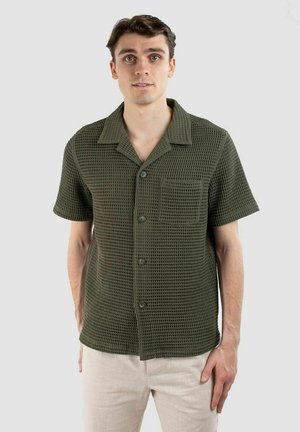 Chemise à manches courtes, de couleur olive, avec un tissu texturé et un motif à carreaux. Présente une poche poitrine et un col classique.