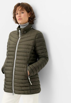 Woman wearing an olive green quilted jacket with front zip and hands in pockets, standing against a white wall.