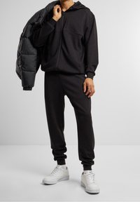 Sudadera negra con cremallera y joggers a juego fabricados en un tejido suave, con puños acanalados y un ajuste relajado, combinados con zapatillas deportivas blancas.