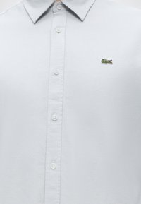 Chemise blanche boutonnée avec col pointu et petit logo crocodile vert brodé sur la poitrine gauche.