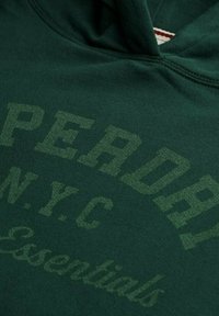 Sudadera verde con capucha hecha de un tejido suave, que presenta un estampado texturizado que dice "SUPERDRY N.Y.C. Essentials" en un tono ligeramente contrastante.