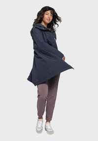 Marineblaue, wasserdichte Jacke mit Kapuze, langen Ärmeln und seitlichen Schlitzen, kombiniert mit hellbraunen Jogginghosen und blauen Turnschuhen mit weißen Akzenten.