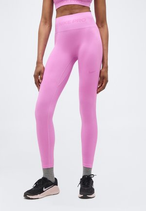Ženska, oblečena v roza Nike Pro legice z visokim pasom, sive nogavice in črne Nike tekaške čevlje, stoji z nekoliko razmaknjenimi nogami.