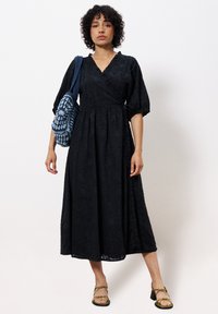 Schwarzes, knielanges Wickelkleid mit puffärmeln, strukturiertem Blumenmuster und gerafftem Taillenbund. Kombiniert mit einer blauen gehäkelten Tasche und Sandalen.