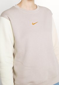 Světle béžová mikina s krémovými rukávy, kulatým výstřihem a jemným logem Nike v oranžové. Měkká textura, ležérní střih a žebrovaný lem.