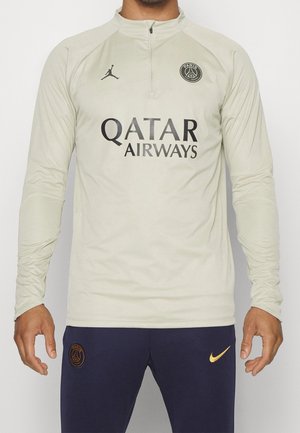 Mężczyzna w jasnobeżowej, długorękawowej bluzie treningowej Paris Saint-Germain z logo Qatar Airways oraz ciemnych spodniach z logo Nike i PSG.