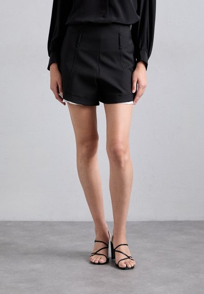 Shorts noirs au design ajusté, avec deux poches avant et une texture lisse. Associés à des talons noirs à brides.