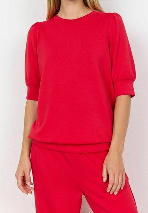 Frau trägt ein leuchtend rotes kurzes Oberteil mit Puffärmeln und passende rote Hosen, stehend vor einem weißen Hintergrund.