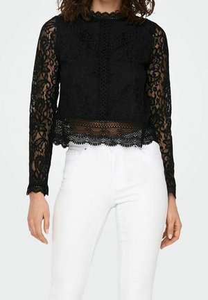 Woman wearing a black long-sleeve lace top with floral patterns and white high-waist pants, standing against a plain background.