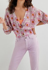 Cardigan en maille avec motif de marguerites roses et oranges, col en V, manches trois-quarts et boutons. Associé à un jean violet clair.