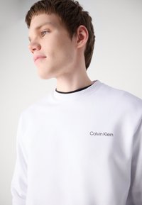 Sudadera blanca hecha de un tejido suave, con cuello redondo y un pequeño logotipo negro de "Calvin Klein" en el pecho. Textura lisa.