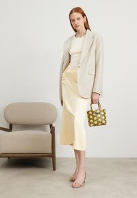 Beige blazer over en hvit topp, gul sateng midi-skjørt, og grønn vevd håndveske. Sandaler med klare hæler. Minimalistisk møbler i bakgrunnen.